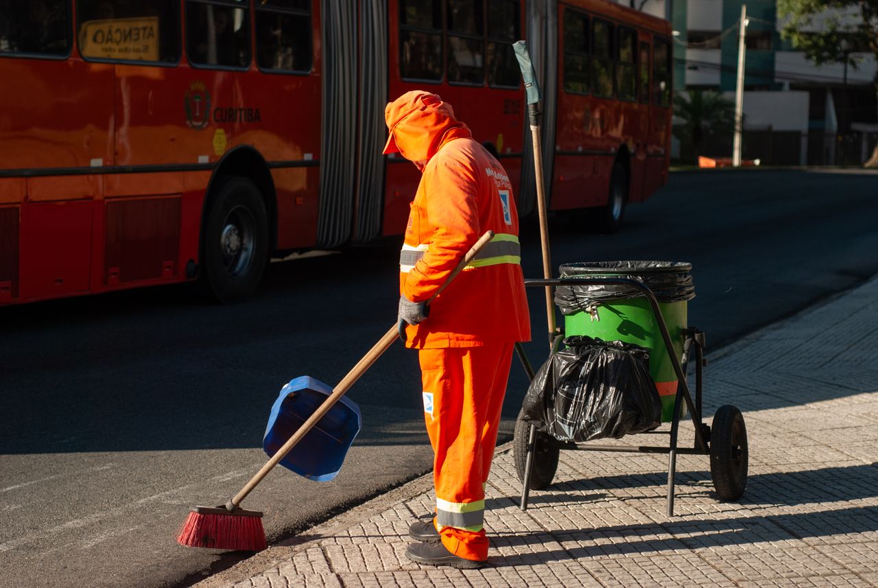 our-story Street cleaner in bright orange uniform sweeping sidewalk near public bus in city setting.